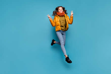 Full body photo of amazing model lady jumping high excited showing v-sign symbol wear stylish windbreaker jeans scarf sweater isolated blue color backgroundの写真素材