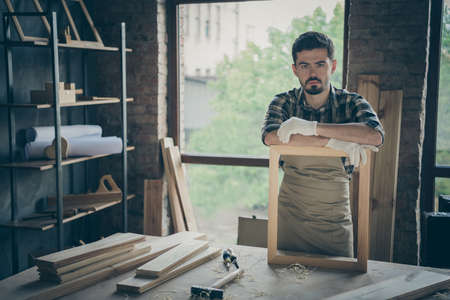 Photo of serious confident joiner in gloves uniform leaning on handmade frame looking at you pensively with planks and tools on canvas tableの写真素材