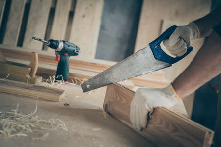 Cropped close up photo of man cutting off unneeded material from wooden block using saw blade in glovesの写真素材