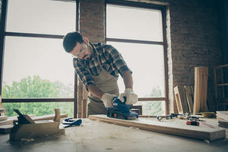 Photo of pensive man wearing checkered shirt eye glasses protecting him from sawdust working by using sanderの写真素材