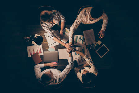 High angle above view photo of four business partners sitting opposite table working together late night holding arms pile stack union formalwear lamp light indoorsの写真素材