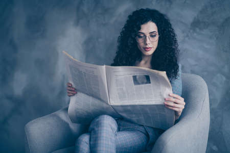 Portrait of her she nice-looking attractive lovely cute charming calm focused peaceful wavy-haired girl sitting in chair reading digest news magazine isolated over gray concrete wall backgroundの写真素材
