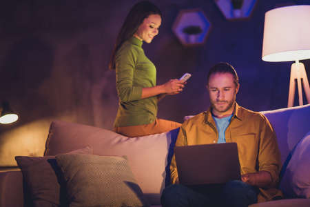 Nice attractive lovely cheerful cheery married spouses sitting on divan using gadget spending free time guy working remotely on start-up at night home house apartmentの写真素材