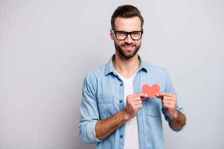Photo of attractive handsome guy holding little red paper heart overjoyed symbolizing cardiology healthcare wear specs casual denim outfit isolated grey color backgroundの写真素材