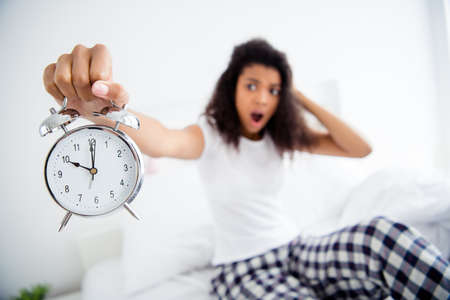Oh no its 10 oclock. Profile photo of crazy mad curly dark skin lady sitting bed sheets holding metal alarm clock morning late work oversleep afraid be fired wear pajama room indoorsの写真素材