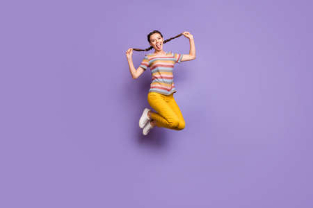 Full length photo of funny lady jumping high summer mood holding pretty braids sticking tongue out mouth wear casual striped t-shirt yellow pants isolated purple color backgroundの写真素材