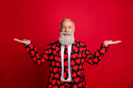 Photo of funky aged handsome man amour role holding open palms offer two sale products demonstrating novelty wear hipster hearts pattern suit blazer tie isolated red color backgroundの写真素材