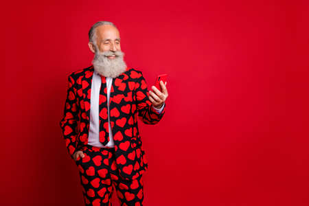 Photo of funny mature guy holding telephone chatting girlfriend write love sms letter wear stylish hearts pattern suit blazer shirt tie pants isolated red color backgroundの写真素材