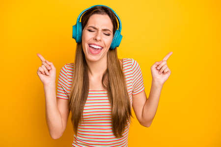 Photo of beautiful lady good mood listen music in cool wireless earphones signing favorite song eyes closed wear casual striped t-shirt isolated yellow color backgroundの写真素材