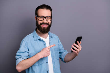 Portrait of positive cheerful guy modern technology shop worker present new smartphone novelty point index finger wear good look clothes isolated over grey color backgroundの写真素材