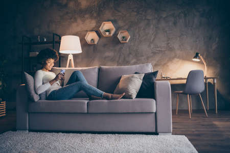 Profile photo of pretty dark skin curly lady domestic atmosphere texting telephone lying comfy couch in lamp light wear casual outfit evening living room indoorsの写真素材
