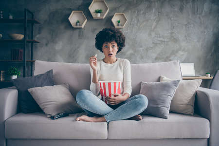 Photo of pretty dark skin wavy lady eating popcorn watching frightening horror film open mouth scary moment sitting cozy couch casual sweater jeans outfit flat indoorsの写真素材