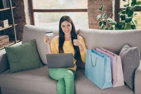Portrait of positive girl student use computer plastic card to pay shopping purchase recommend show like sign sit cozy couch in house indoorsの写真素材