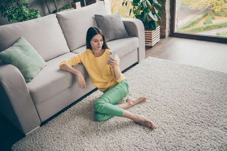 Top above high angle photo of positive cheerful girl sit floor carpet barefoot use smartphone read social network news wear yellow jumper green pants trousers in house indoorsの写真素材