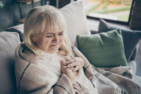 Photo of unhappy white haired aged granny holding chest zone cardiac difficulties afraid heart attack sitting sofa divan covered plaid blanket living room indoorsの写真素材