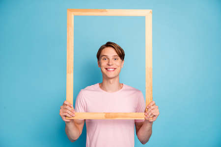Portrait of funky cheerful guy hold wooden brick frame make picture of himself wear casual style clothing isolated over blue color backgroundの写真素材