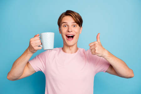 Portrait of amazed funky hipster guy drink hot caffeine beverage hold cup like show thumb up sign scream wear stylish pink t-shirt isolated over blue color backgroundの写真素材