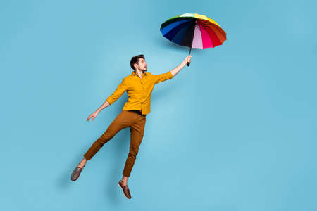 Full length photo of crazy guy jumping high holding colored bright umbrella, flying up higher wear yellow shirt trousers isolated blue color backgroundの写真素材
