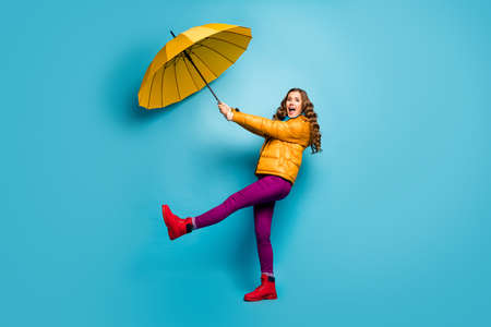 Help me. Full size profile photo of crazy shocked lady hold umbrella flying up with wind blowing wear yellow overcoat scarf violet pants red footwear isolated blue color backgroundの写真素材