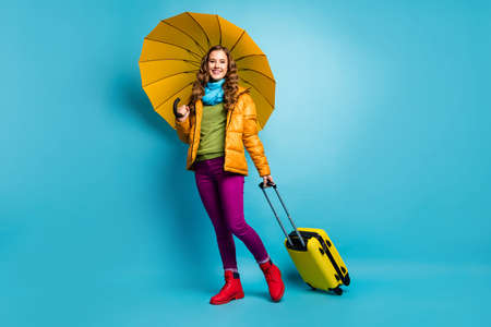Full body photo of beautiful traveler lady hold umbrella rolling suitcase walk airport registration wear yellow overcoat blue scarf trousers boots isolated blue color backgroundの写真素材