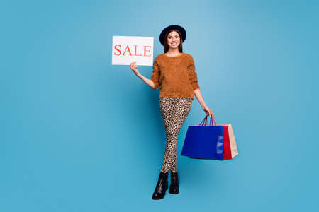 Fall season off sales. Full length photo of addicted girl hold bags show text word sale recommend retail offer wear brown sweater trousers vintage headwear isolated blue color backgroundの写真素材