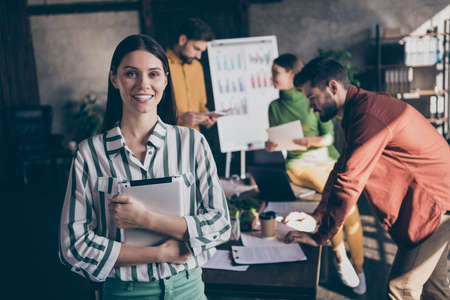 Photo of cheerful positive pretty cute entrepreneur holding electronic device smile toothy standing on background of her team developing projectの写真素材