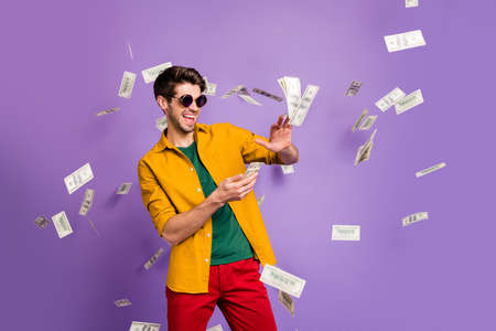 Photo of cheerful excited white trendy man throwing cash away showing his wealthiness smiling toothily in red pants isolated violet color backgroundの写真素材