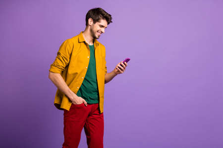 Photo of cheerful man holding hand in pocket of red pants smiling toothily holding telephone browsing isolated pastel violet color backgroundの写真素材