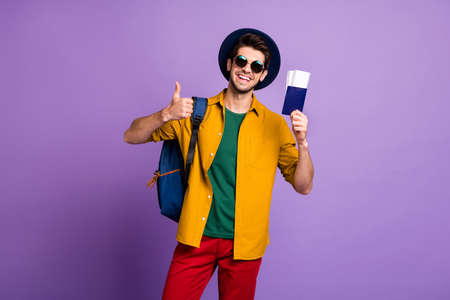 Positive guy hold airport tickets go weekends show thumb-up advise choose first-class trip wear yellow shirt red pants trousers backpack blue headwear isolated violet color backgroundの写真素材