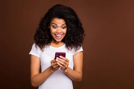 Portrait of amazed crazy afro american girl blogger use cellphone get social network notification look scream wow omg wear white t-shirt isolated over brown color backgroundの写真素材