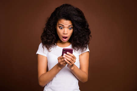 Portrait of crazy funky afro american girl use smart phone read social network information impressed scream wow omg wear casual style clothing isolated over brown color backgroundの写真素材