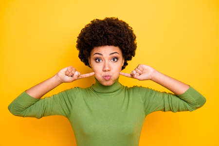 Photo of stylish cheerful girl blowing her cheeks with forefingers touching them looking at you with lips pouted fooling isolated vivid color backgroundの写真素材