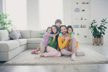 Portrait of nice attractive adorable careful cheerful cheery family pre-teen kids mom dad sitting on carpet floor embracing at cozy comfortable light white interior style house apartment living-roomの写真素材