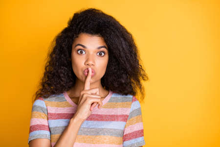 Close-up portrait of her she nice attractive attentive mysterious wavy-haired girl in striped t-shirt showing shh sign isolated on bright vivid shine vibrant yellow color backgroundの写真素材