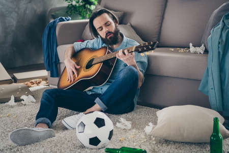 Photo of hipster guy with long beard sitting carpet near sofa holding guitar dont mind chaos after stag party messy dirty flat singing songs careless person indoorsの写真素材