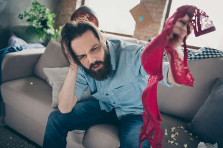 Photo of hipster crazy guy sitting sofa holding head looking red brassiere not remember night intoxicated had stag party suffer hangover morning headache messy flat indoorsの写真素材