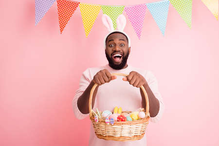 Photo of funny dark skin guy came, easter party hold festive basket ginger bread painted eggs cookies wear sweater bunny ears colorful flags hang pastel pink color backgroundの写真素材
