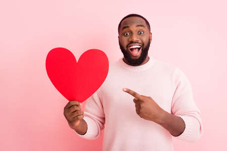 Photo of attractive nice dark skin guy hold red paper heart boyfriend, directing finger presenting funny postcard girlfriend wear sweater isolated pastel pink color backgroundの写真素材