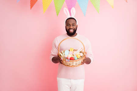 Photo of funny dark skin guy meet guests easter party hold festive, basket ginger bread cookies paint eggs wear sweater pants bunny ears colorful flags hang pink color backgroundの写真素材