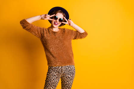 Photo of beautiful pretty funny lady hold fingers v-sign symbol near eyes good mood wear fluffy sweater leopard pants sun specs retro cap isolated yellow color backgroundの写真素材