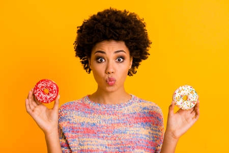Portrait of funny dreamy afro american girl hold two donuts send air kisses enjoy valentine day snack nutrition wear colorful shine jumper isolated over yellow color backgroundの写真素材