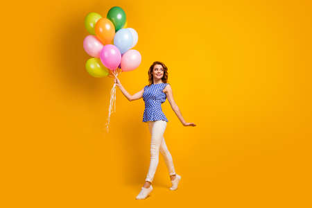 Full body profile photo of funny foxy lady hold many colorful balloons walk street festive mood party wear polka-dot blue shirt white pants shoes isolated yellow color backgroundの写真素材
