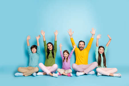 Nice attractive ecstatic cheerful cheery big full family pre-teen kids mom dad sitting in lotus pose enjoying celebrating rising hands up isolated on bright vivid shine vibrant blue color backgroundの写真素材