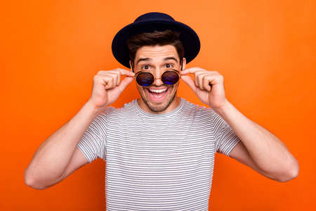 Photo of attractive crazy funky youngster guy taking off cool eyeglasses see advert banner wear retro hat sun specs striped t-shirt isolated bright orange color backgroundの写真素材