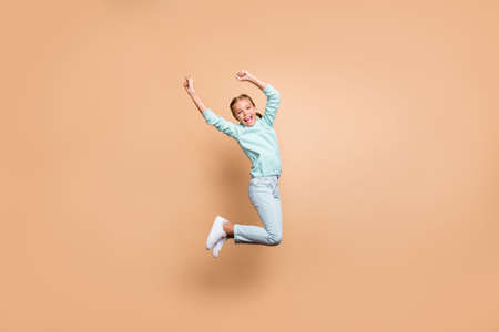Full body profile photo of beautiful funny little lady jumping high up cheerleader celebrating winning raise fists wear blue sweater jeans footwear isolated beige color backgroundの写真素材