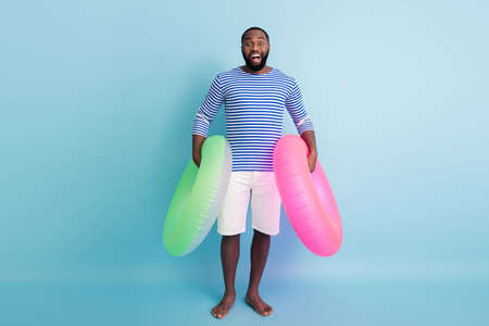 Full length photo of funny dark skin guy holding two green pink life buoy ready to swim ocean sea...