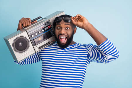 Photo of funny cool dark skin guy open mouth hold retro cassette recorder on shoulder students party chill wear sun glasses striped sailor shirt isolated blue color backgroundの写真素材