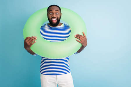 Photo of cheerful dark skin guy hold green life buoy around neck ready swim ocean sea traveler good mood sunny day wear striped sailor shirt isolated blue color backgroundの写真素材
