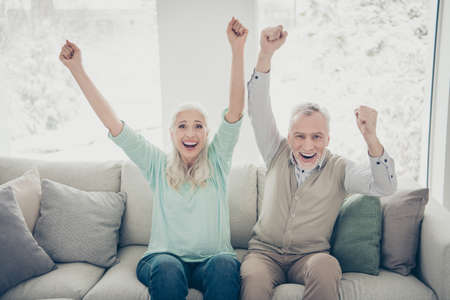 Portrait of cheerful married people in teal brown pullovers raising fists screaming yeah sitting divan apartmentの写真素材
