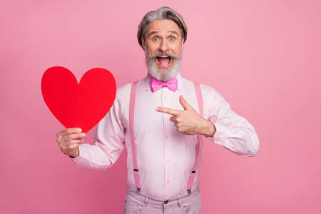 Portrait of his he nice attractive imposing ecstatic cheerful cheery grey-haired man holding in hands demonstrating big large heart soulmate isolated over pink pastel color backgroundの写真素材